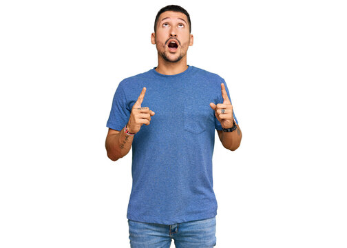 Handsome man with tattoos wearing casual clothes amazed and surprised looking up and pointing with fingers and raised arms.
