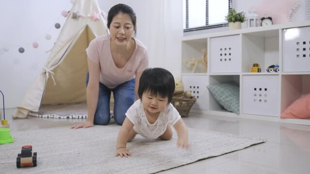 cheerful asian mother praising and applauding for her lovely toddler child who&rsquo;s learning to crawl on the ground in the game room at home.