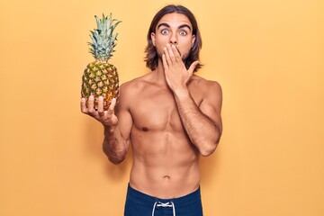 Young handsome man wearing swimwear holding pineapple covering mouth with hand, shocked and afraid...