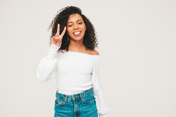 Beautiful black woman with afro curls hairstyle.Smiling model in sweater and trendy jeans clothes. Sexy carefree female posing on white background in studio. Tanned and cheerful.Shows peace sign