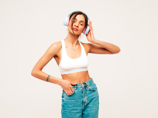 Beautiful smiling woman dressed in jeans clothes. Sexy carefree model listening music in wireless headphones. Adorable and positive female posing on grey background in studio. Closed eyes