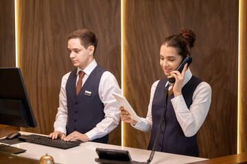 One of two young hotel receptionists standing by counter, looking at touchpad display and...