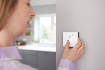 Woman At Home In Kitchen Turning Down Central Heating Thermostat To save Energy And Money
