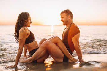 Happy young fit couple sit on beacjin the sea or ocean and embrace each other with love at summer sunset. Online dating, summer love, vacation concept.