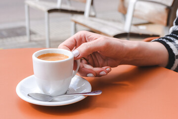 hand of a caucasian young woman holding a white cup of coffee on the top of an orange table at a cafe terrace