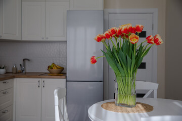 A bouquet of bright tulips on the table. A bright doll in the interior of a light kitchen. Spring flowers. Fresh orange buds. Cozy kitchen