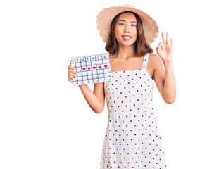 Obraz na płótnie Canvas Young beautiful chinese girl wearing summer hat holding heart calendar doing ok sign with fingers, smiling friendly gesturing excellent symbol