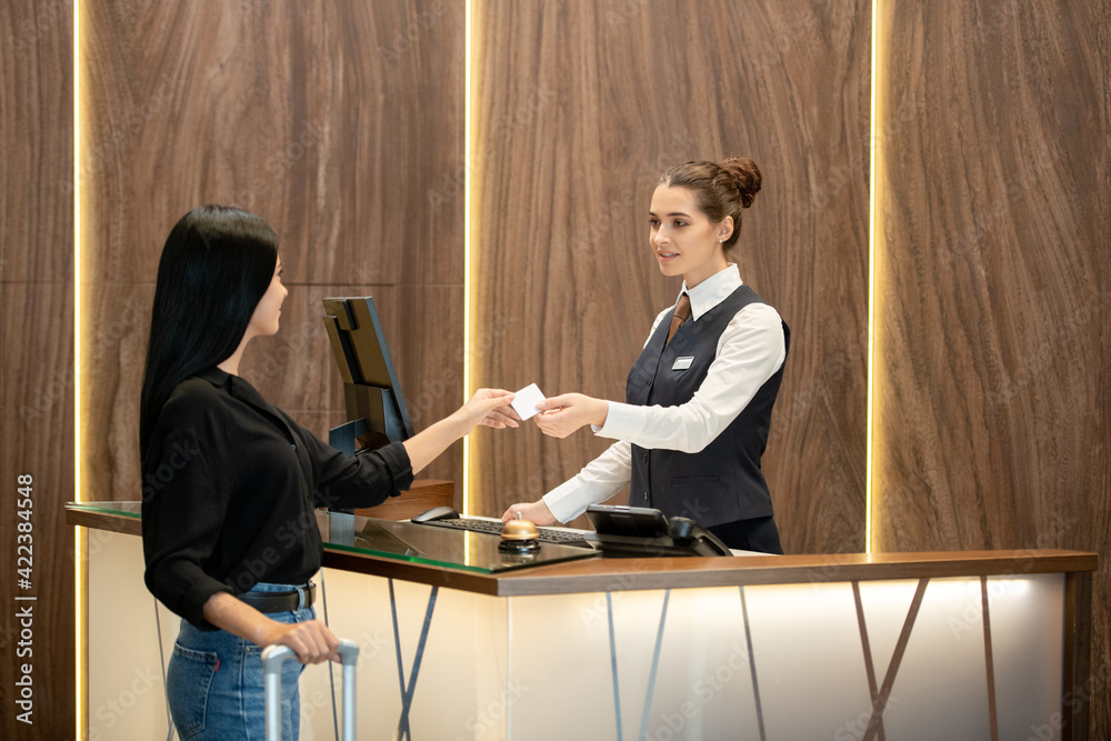 Poster young brunette receptionist in uniform standing by workplace and passing plastic card to one of gues