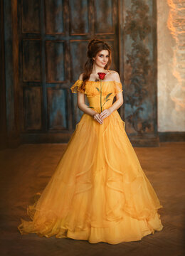 Girl beauty fantasy princess in yellow long historical, medieval silk dress holding flower red rose in her hands. Background of old gothic castle room. Fairy tale bewitched queen. Happy woman smiles