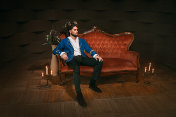 Strong handsome confident man with ram horns on head, sitting on red vintage sofa in dark room....