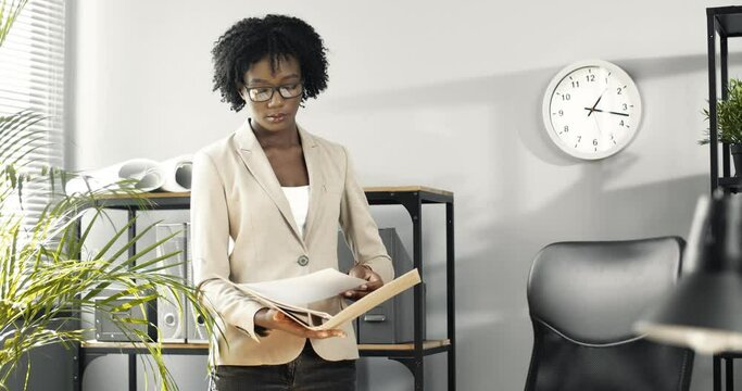 Smart successful business lady in glasses with curly hair stands in modern office and browses documents in folder.