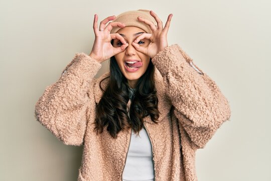 Young hispanic woman wearing wool sweater and winter hat doing ok gesture like binoculars sticking tongue out, eyes looking through fingers. crazy expression.