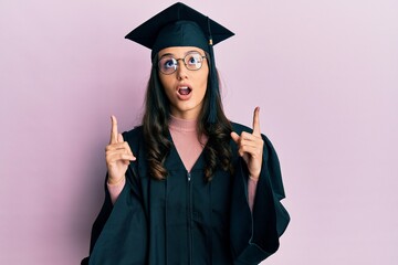 Young hispanic woman wearing graduation cap and ceremony robe amazed and surprised looking up and pointing with fingers and raised arms.