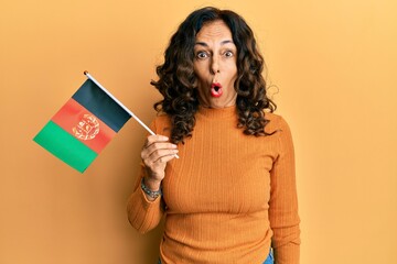 Middle age hispanic woman holding afghanistan flag scared and amazed with open mouth for surprise, disbelief face