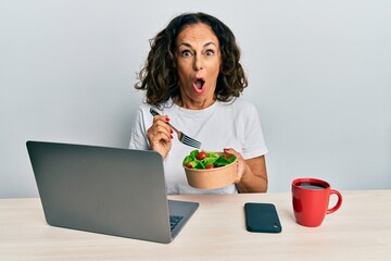 Beautiful middle age woman working at the office eating healthy salad afraid and shocked with...