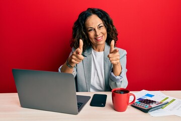 Beautiful middle age woman working at the office drinking a cup of coffee pointing fingers to...