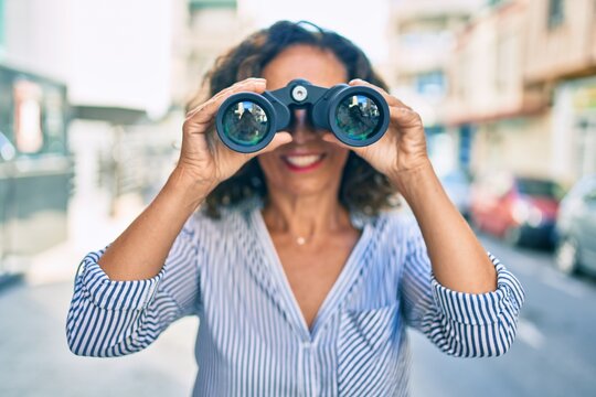 Middle age hispanic woman smiling happy looking for new opportunity using binoculars at the city.