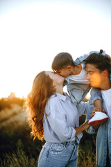 happy young family in the park at sunset. happy boy sits on dad's shoulders. mom kisses her son. sun rays on faces. young stylish family in casual outdoor clothes
