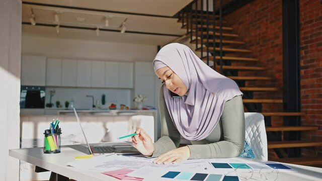 Portrait of modern Muslim woman working remotely at home. There is a laptop and documents on the table. The girl wears a hijab, talks on the phone and makes notes with a pencil