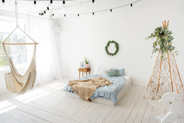 Stylish, trendy interior in Scandinavian style. In white loft room, there is wigwan macrame with green ivy, bed on the floor, wooden rocking horse, hand-made hammock hanging by the window. Copy space