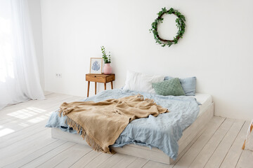 Stylish, trendy interior in Scandinavian style. In a white loft room, a bed on the floor with a blue bed, brown rug, a wicker wreath on the wall with ivy, wooden bedside table with a vase. Copy space