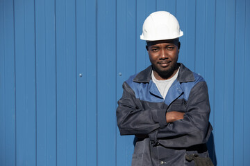 Happy young factory worker of African ethnicity in workwear and hardhat crossing his arms by chest while standing in front of camera outdoors