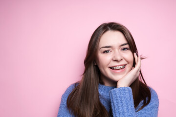 Portrait of an attractive brunette girl in braces, in a blue sweater smiling on a pink background....