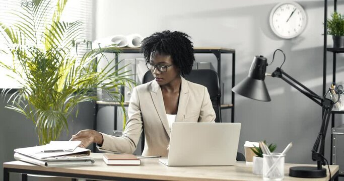 Smart African-American businesswoman with glasses and curly hair is sitting at table in modern office, working on laptop and comparing information with papers.