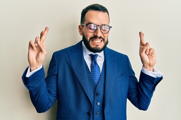 Young man with beard wearing business suit and tie gesturing finger crossed smiling with hope and eyes closed. luck and superstitious concept.