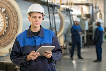 Young female factory worker in overalls and hardhat looking at you while standing in front of camera in workshop and using digital tablet