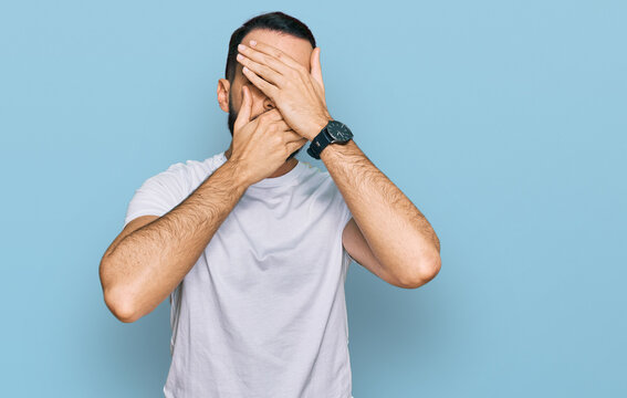 Young man with beard wearing casual white t shirt covering eyes and mouth with hands, surprised and shocked. hiding emotion