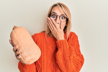Beautiful blonde woman holding healthy fresh pumpkin covering mouth with hand, shocked and afraid...