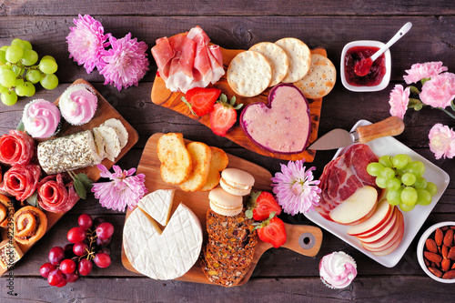 Mother's Day theme charcuterie table scene against a dark wood background. Assorted cheese, meat, fruit and sweet appetizers. Top down view.