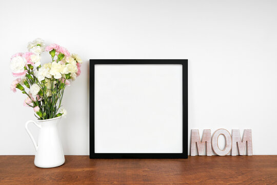 Mock up black frame with vase of carnation flowers and wooden MOM letters. Mother's Day decor theme. Wood shelf against a white wall. Copy space.