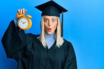Beautiful blonde woman wearing graduation cap and ceremony robe holding alarm clock scared and...