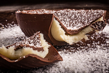 Close up of a cracked chocolate easter egg with coconut filling and grated coconut on the top on a wooden table.
