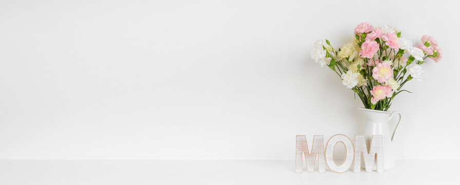 Mother's Day decor with vase of carnation flowers and wooden MOM letters on a white shelf against a white wall banner background. Copy space.