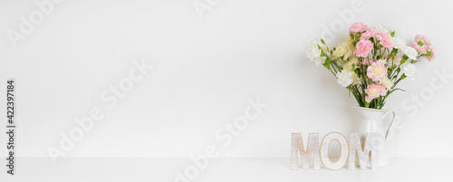 Mother's Day decor with vase of carnation flowers and wooden MOM letters on a white shelf against a white wall banner background. Copy space.