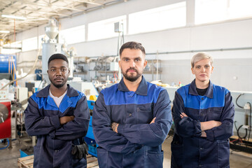 Three young intercultural workers of modern plant in overalls standing in front of camera against industrial machines in large workshop