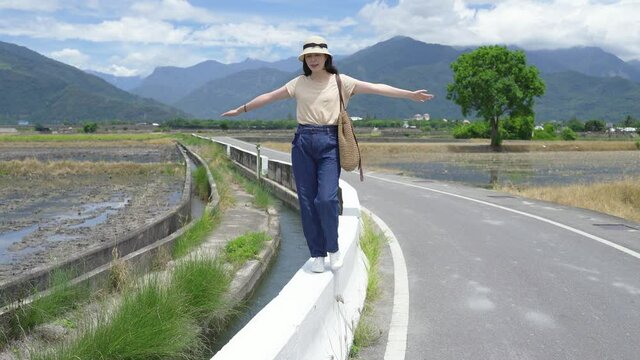 woman with extended arms is balancing on the white stone pier beside a rural path with a curve and scenic landscape on a bright summer&rsquo;s day, full length view.