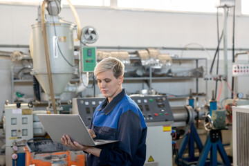 Young serious female worker in overalls looking at laptop display while looking through online technical data against industrial machines