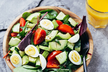 Green salad with tomatoes, avocado, cucumber and eggs on wooden plate and orange juice on the table.