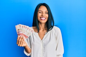 Young african american woman holding 10 colombian pesos banknotes winking looking at the camera with sexy expression, cheerful and happy face.