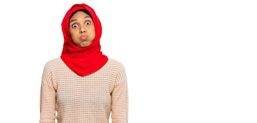 Young african american woman wearing traditional islamic hijab scarf puffing cheeks with funny face. mouth inflated with air, crazy expression.
