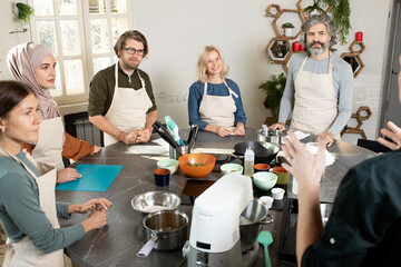 Group of young and mature people in aprons looking at male cooking coach and listening to him during master class in the kitchen