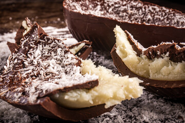 Close up of a cracked chocolate easter egg with coconut filling and grated coconut on the top on a wooden table.