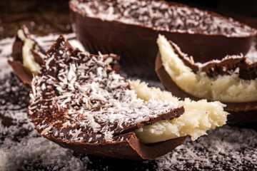 Close up of a cracked chocolate easter egg with coconut filling and grated coconut on the top on a wooden table.