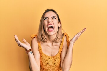 Young caucasian blonde woman wearing casual yellow t shirt crazy and mad shouting and yelling with aggressive expression and arms raised. frustration concept.