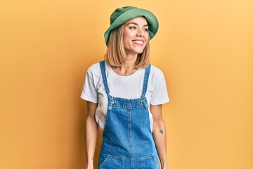 Young caucasian blonde woman wearing denim jumpsuit and hat with 90s style looking away to side...