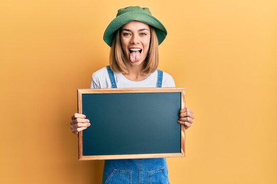 Young caucasian blonde woman holding blackboard sticking tongue out happy with funny expression.